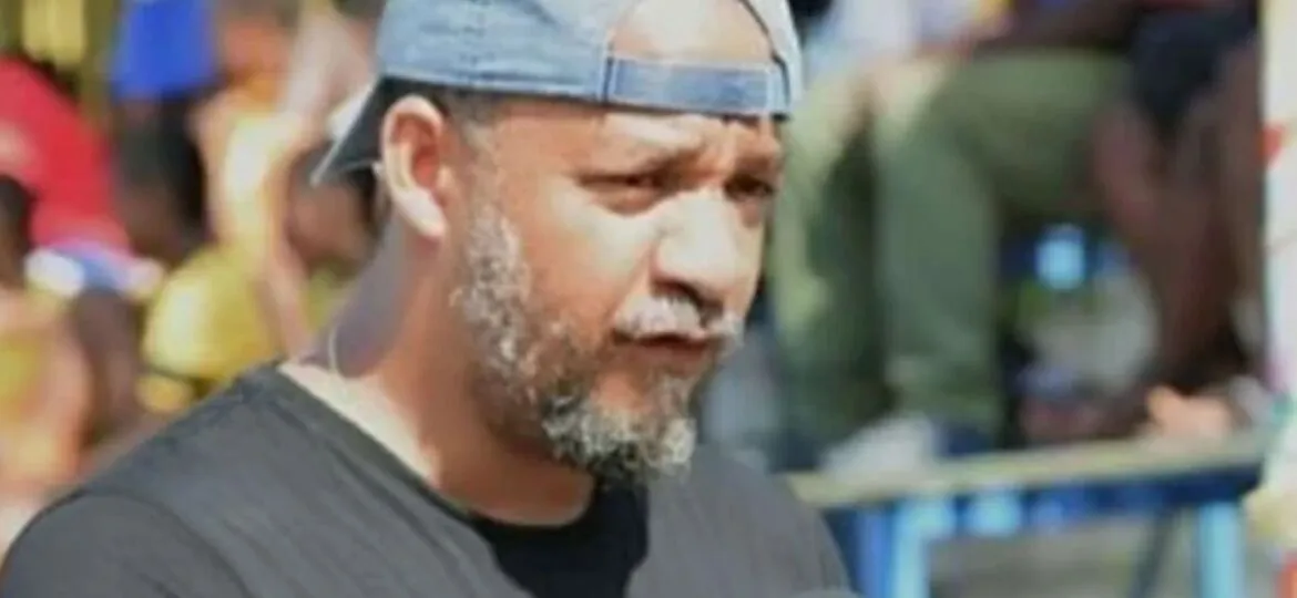 Kako Bourjolly speaking outdoors, wearing a black shirt and a backward baseball cap, with a blurred crowd in the background.